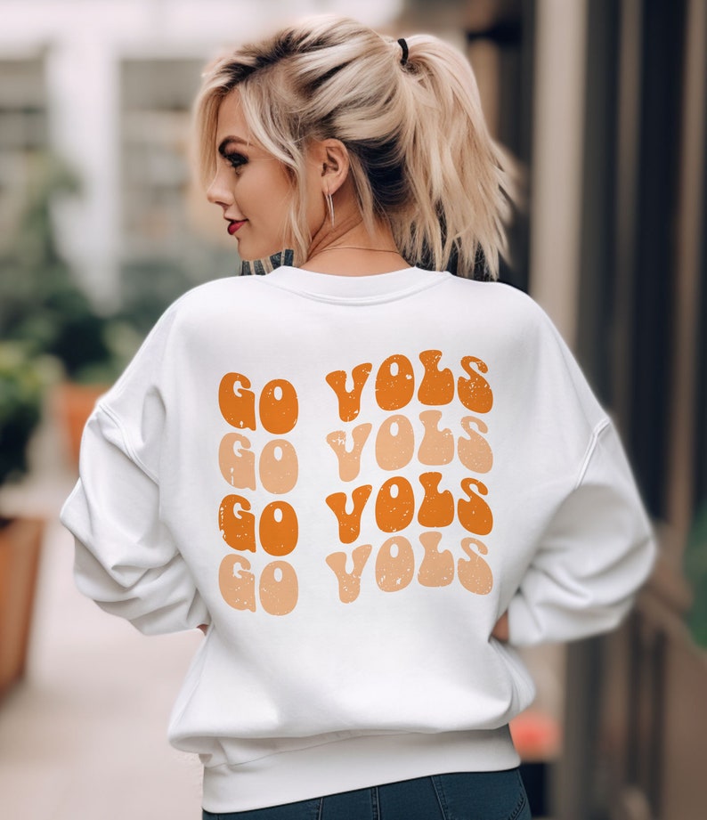 K&ouml;nnte beinhalten: Wei&szlig;er Pullover mit dem Text "GO VOLS", der f&uuml;nfmal in einer Retro-Schriftart in Orange und Pfirsich wiederholt wird.