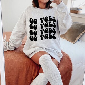 K&ouml;nnte beinhalten: Ein graues Sweatshirt mit dem Text "GO VOLS", der in einer Retro-Schriftart wiederholt wird. Das Sweatshirt wird von einer Person mit wei&szlig;en Kniestr&uuml;mpfen getragen.