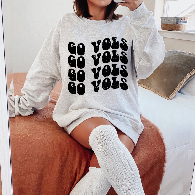 K&ouml;nnte beinhalten: Ein graues Sweatshirt mit dem Text "GO VOLS", der in einer Retro-Schriftart wiederholt wird. Das Sweatshirt wird von einer Person mit wei&szlig;en Kniestr&uuml;mpfen getragen.
