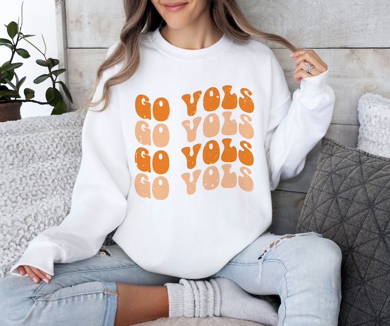 K&ouml;nnte beinhalten: Wei&szlig;er Pullover mit dem Text "Go Vols", der viermal in einem Retro-Stil in Orange und Pfirsich wiederholt wird.