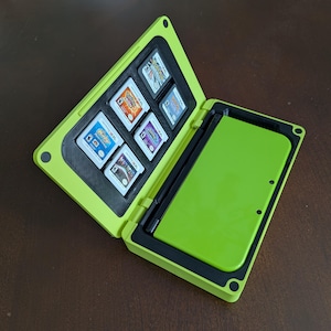 May include: A lime green hinged storage case featuring a black interior with dedicated slots for five small rectangular game cartridges and a larger rectangular compartment designed to hold a handheld gaming console securely in place.