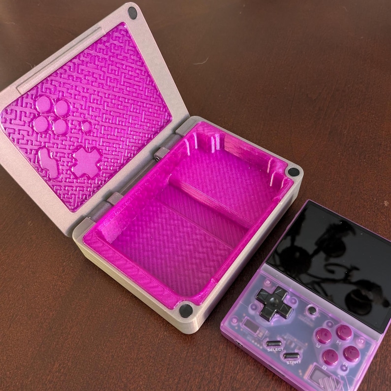 May include: A purple and grey 3D printed case with a textured lid, open to reveal a purple interior. The case is designed to hold a small handheld gaming device with a black screen, purple buttons, and a black directional pad.