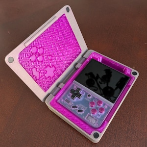 May include: A pink and grey handheld gaming console with a screen, buttons, and a directional pad. The console is open and the screen is showing a reflection of the surrounding environment.