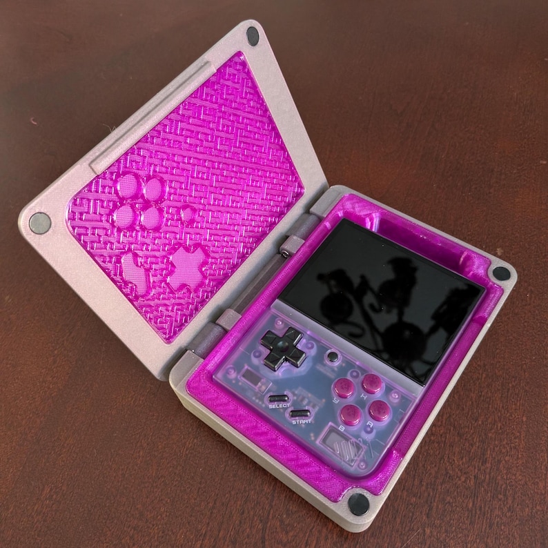 May include: A pink and grey handheld gaming console with a screen, buttons, and a directional pad. The console is open and the screen is showing a reflection of the surrounding environment.