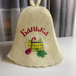 May include: A light beige sauna hat with the embroidered word "Банька" in red Cyrillic script. The hat features a colorful embroidered design of a sauna bucket, ladle, and a sprig of greenery.