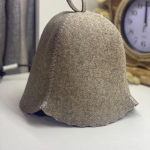 Könnte beinhalten: Hellbrauner Sauna-Hut aus Filz mit rundem Oberteil und kleiner Schlaufe zum Aufhängen. Der Hut hat einen gewellten Rand und ist mit weißem Faden genäht. Er schützt den Kopf vor Hitze in der Sauna.