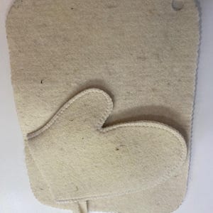 Puede incluir: Una alfombrilla rectangular de fieltro beige con esquinas redondeadas y un pequeño orificio circular en la esquina superior derecha, con una pieza de fieltro beige más pequeña en forma de manopla superpuesta con bordes cosidos.