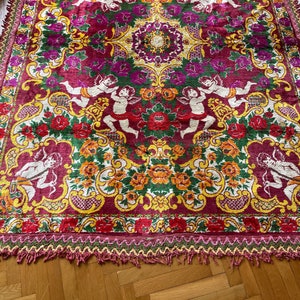Puede incluir: Una alfombra grande y ornamentada con un fondo burdeos intenso. La alfombra presenta un patr&oacute;n repetido de querubines, flores y detalles dorados. Los bordes est&aacute;n adornados con flecos.