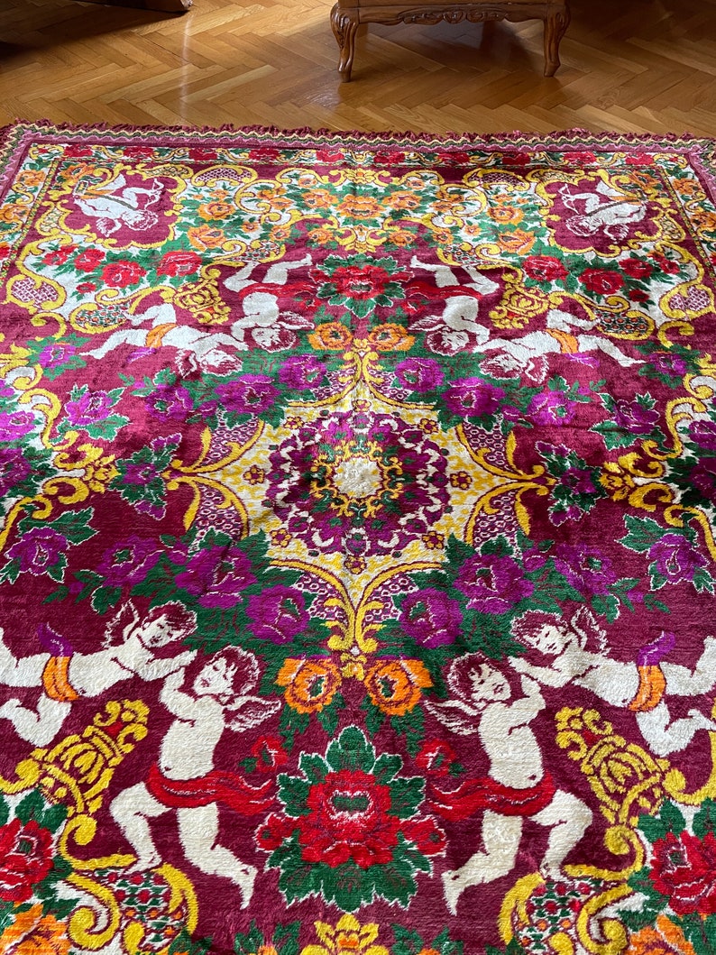 Puede incluir: Una alfombra grande y ornamentada con un dise&ntilde;o rico y detallado. La alfombra presenta un fondo burdeos oscuro con un patr&oacute;n repetido de querubines, flores y vides en tonos de rojo, verde, amarillo y morado. La alfombra tiene un borde con flecos.