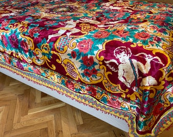 Puede incluir: Una manta de terciopelo vintage con un diseño floral y de querubines colorido. La manta tiene un borde con flecos y está en tonos de rojo, amarillo, verde y blanco.