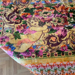 May include: A close-up of a vintage rug with a floral design. The rug has a yellow background with a pattern of flowers in pink, purple, orange, and green. A cherub figure is also visible.