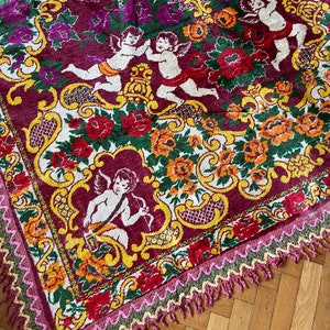 Puede incluir: Textil decorativo con un patr&oacute;n repetido de querubines, flores y adornos ornamentados en tonos burdeos, naranja, amarillo y verde. Los bordes est&aacute;n acabados con un ribete en zigzag rosa y verde y flecos.