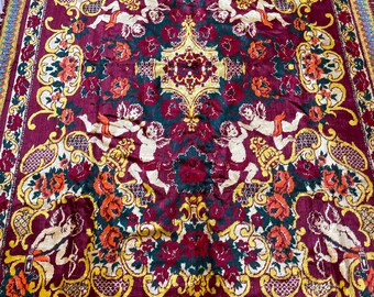 Puede incluir: Una alfombra vintage roja, amarilla y verde con un diseño floral y figuras de querubines. La alfombra tiene un borde de flecos.