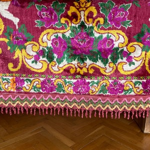 Puede incluir: Primer plano de un textil burdeos con un estampado floral. El dise&ntilde;o presenta grandes flores moradas, hojas verdes y adornos amarillos sobre un fondo blanco. Se aprecia un borde decorativo con flecos.