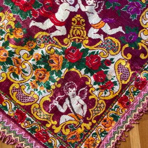 Puede incluir: Textil decorativo con fondo burdeos adornado con motivos florales y querubines. El dise&ntilde;o incluye rosas rojas, amarillas y naranjas, y un borde con flecos rosas y verdes. Los querubines son blancos.