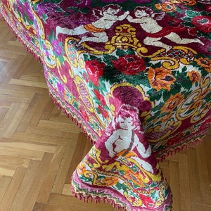 Puede incluir: Un textil ricamente estampado, probablemente un mantel o tela decorativa, con un complejo dise&ntilde;o floral y de querubines. La paleta de colores incluye rojos intensos, morados, amarillos y verdes. Los bordes est&aacute;n acabados con flecos. La tela es probablemente de terciopelo.