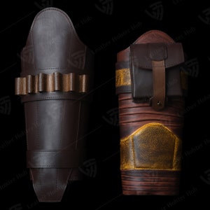 Op de afbeelding: Twee bruine leren armbeschermers met decoratieve stiksels. Een beschermer heeft een klein zakje aan de zijkant. Beide beschermers hebben een bruine leren band met decoratieve stiksels.