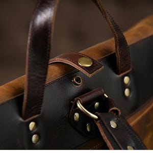 Op de afbeelding: Close-up van een bruine en zwarte leren rugzak. De tas heeft donkerbruine leren riemen, messingkleurige klinknagels en een gesp. De tas heeft een distressed, vintage look.