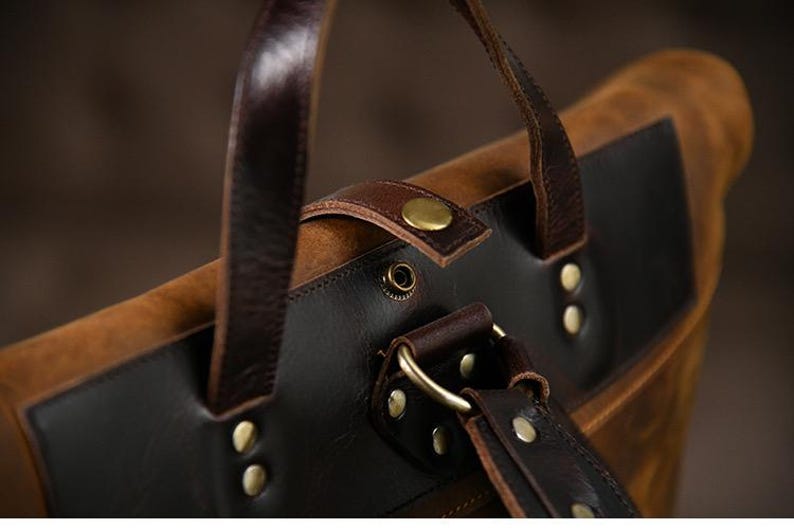 Op de afbeelding: Close-up van een bruine en zwarte leren rugzak. De tas heeft donkerbruine leren riemen, messingkleurige klinknagels en een gesp. De tas heeft een distressed, vintage look.