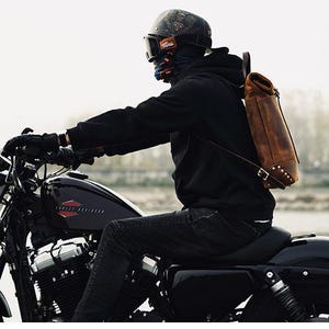 Op de afbeelding: Een persoon rijdt op een zwarte Harley-Davidson motorfiets, gekleed in een zwarte hoodie, helm en bruine leren rugzak. De motorfiets heeft een zwart zadel en chromen accenten. De achtergrond is wazig, wat een buitenomgeving suggereert.