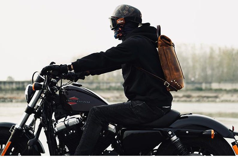 Op de afbeelding: Een persoon rijdt op een zwarte Harley-Davidson motorfiets, gekleed in een zwarte hoodie, helm en bruine leren rugzak. De motorfiets heeft een zwart zadel en chromen accenten. De achtergrond is wazig, wat een buitenomgeving suggereert.
