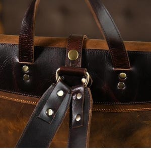 Op de afbeelding: Close-up van een bruine leren rugzak met donkerbruine accenten. De tas heeft een klepsluiting met een gesp en verstelbare riemen. Het leer heeft een distressed look en de hardware is goudkleurig.