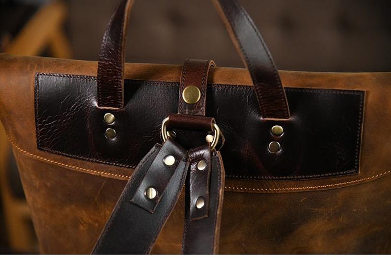 Op de afbeelding: Close-up van een bruine leren rugzak met donkerbruine accenten. De tas heeft een klepsluiting met een gesp en verstelbare riemen. Het leer heeft een distressed look en de hardware is goudkleurig.