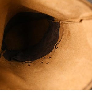 Op de afbeelding: Een close-up van de binnenkant van een lichtbruine leren tas. De binnenkant is donkerbruin, met zichtbare stiksels en klinknagels langs de randen. De opening van de tas is breed en toont de diepte en vorm.