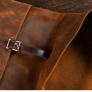 Op de afbeelding: Close-up van een bruine leren tas met een klepsluiting. De tas heeft een donkerbruine leren riem met een goudkleurige gesp. Het leer heeft een verweerde, getextureerde uitstraling.