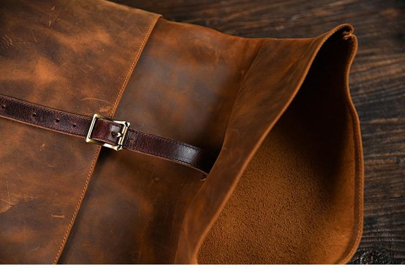 Op de afbeelding: Close-up van een bruine leren tas met een klepsluiting. De tas heeft een donkerbruine leren riem met een goudkleurige gesp. Het leer heeft een verweerde, getextureerde uitstraling.