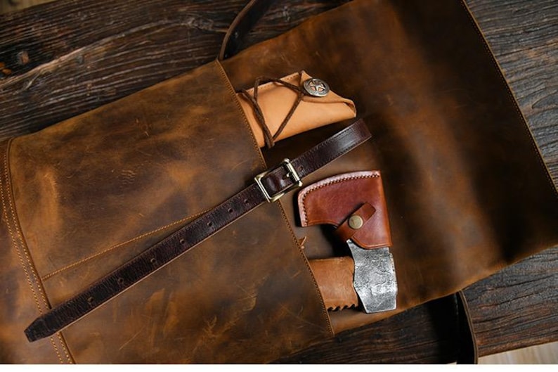 Op de afbeelding: Een bruine leren tas met een donkerbruine riem en een gouden gesp. In de tas zit een klein leren zakje met een zilveren knoop en een leren foedraal met een kleine bijl.