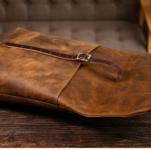 Op de afbeelding: Een bruine leren rugzak met een distressed afwerking. De tas heeft een klepsluiting met een gesp en een bruine leren riem. De tas ligt op een donker houten oppervlak.