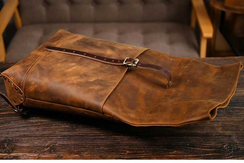 Op de afbeelding: Een bruine leren rugzak met een distressed afwerking. De tas heeft een klepsluiting met een gesp en een bruine leren riem. De tas ligt op een donker houten oppervlak.