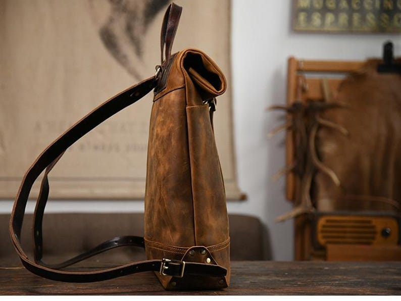 Op de afbeelding: Een bruine leren rugzak met verstelbare riemen en een handvat aan de bovenkant. De tas heeft een oprolbare sluiting en een gesp aan de basis. Het leer heeft een distressed afwerking, waardoor het een vintage uitstraling heeft. De tas staat op een houten oppervlak.