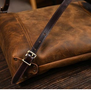 Op de afbeelding: Een close-up van een bruine leren tas met een donkerbruine riem en een goudkleurige gesp. De tas heeft een distressed afwerking en ligt op een houten oppervlak. De riem is met klinknagels aan de tas bevestigd.