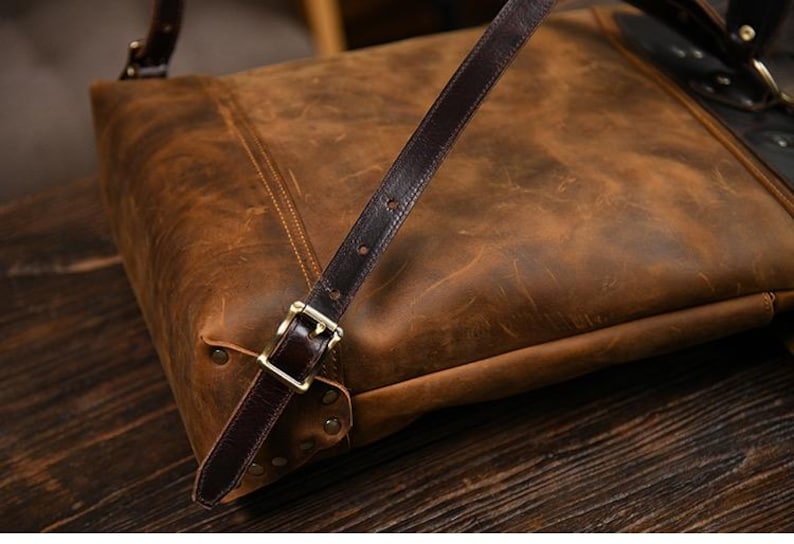 Op de afbeelding: Een close-up van een bruine leren tas met een donkerbruine riem en een goudkleurige gesp. De tas heeft een distressed afwerking en ligt op een houten oppervlak. De riem is met klinknagels aan de tas bevestigd.