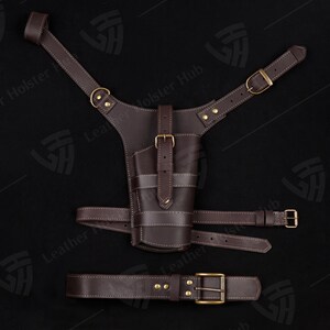 May include: A brown leather holster with a belt and shoulder straps. The holster has a stitched design and brass buckles.