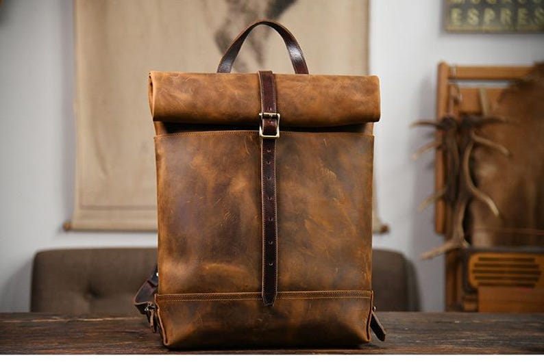 Op de afbeelding: Een bruine leren rugzak met een oprolbare sluiting en een verticale riem die met een gesp is vastgezet. De tas heeft een handvat aan de bovenkant en is gemaakt van distressed leer. De rugzak staat op een houten oppervlak.