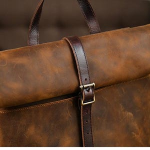 Op de afbeelding: Bruine leren rugzak met een oprolbaar ontwerp. De tas heeft een donkerbruine leren riem met een metalen gespsluiting. De tas heeft een handvat aan de bovenkant en een getextureerde afwerking.
