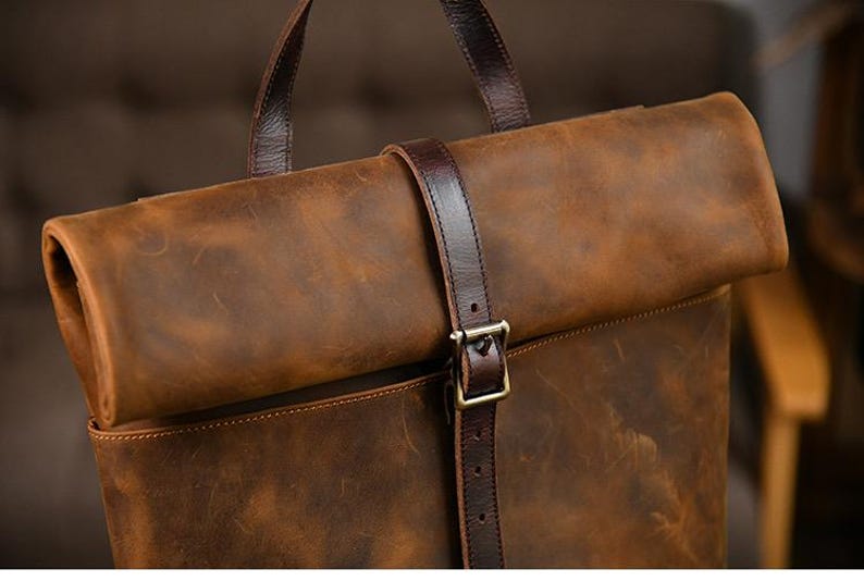 Op de afbeelding: Bruine leren rugzak met een oprolbaar ontwerp. De tas heeft een donkerbruine leren riem met een metalen gespsluiting. De tas heeft een handvat aan de bovenkant en een getextureerde afwerking.