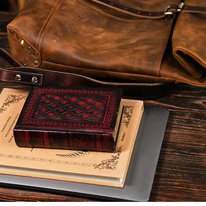 Op de afbeelding: Een bruine leren rugzak met een klepsluiting en riem ligt bovenop een stapel boeken en een kleine, rood-zwarte, lederen doos. De doos heeft een ruitpatroon en ligt bovenop een boek met decoratieve randen.