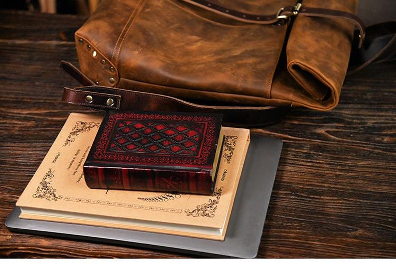 Op de afbeelding: Een bruine leren rugzak met een klepsluiting en riem ligt bovenop een stapel boeken en een kleine, rood-zwarte, lederen doos. De doos heeft een ruitpatroon en ligt bovenop een boek met decoratieve randen.
