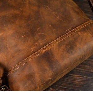 Op de afbeelding: Close-up van een bruine leren rugzak met zichtbare stiksels en een schouderriem. Het leer heeft een distressed look met kleur- en textuurvariaties, wat wijst op een vintage of rustieke stijl. De tas lijkt leeg te zijn.