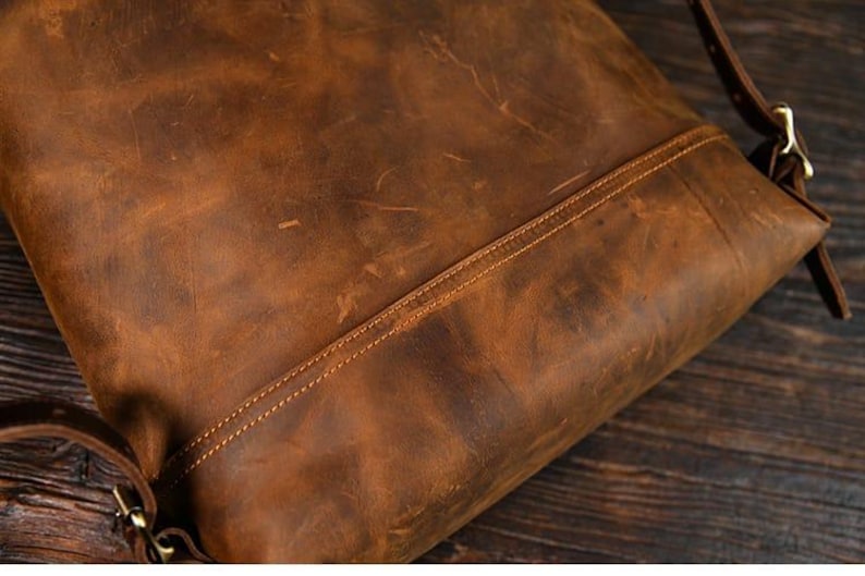 Op de afbeelding: Close-up van een bruine leren rugzak met zichtbare stiksels en een schouderriem. Het leer heeft een distressed look met kleur- en textuurvariaties, wat wijst op een vintage of rustieke stijl. De tas lijkt leeg te zijn.