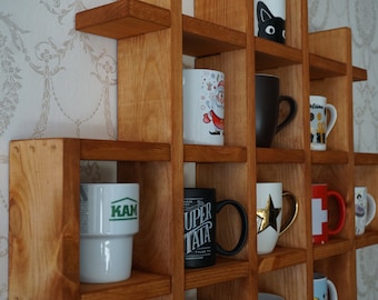 Może przedstawiać: Drewniany regał kratowy z wieloma kwadratowymi przegródkami mieszczącymi różne ceramiczne kubki. Kubki mają różnorodne wzory, w tym czarnego kota, ilustrację Świętego Mikołaja, złotą gwiazdę, czerwoną flagę Szwajcarii oraz napisy KAM i SUPER TATA.