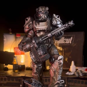 May include: A detailed figurine of a character from the Fallout video game series, clad in metallic armor and wielding a large weapon. The base features a copper-toned relief with stars and a hazard symbol. The word "Fallout" is visible.