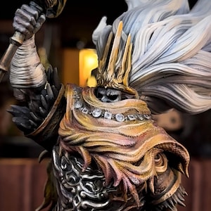 May include: A detailed figurine of a warrior in ornate armor, featuring gold, bronze, and blue hues. The figure has a crown and flowing white hair. The warrior is holding a weapon aloft, showcasing intricate design and craftsmanship.