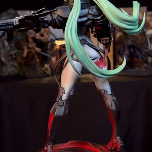 K&ouml;nnte beinhalten: Detaillierte Figur einer weiblichen Figur in einem futuristischen Outfit, die eine Waffe h&auml;lt. Die Figur hat langes, flie&szlig;endes, mintgr&uuml;nes Haar und ist auf einer kreisf&ouml;rmigen Basis mit roter Beleuchtung positioniert. Das Outfit ist haupts&auml;chlich schwarz und grau mit roten Akzenten.