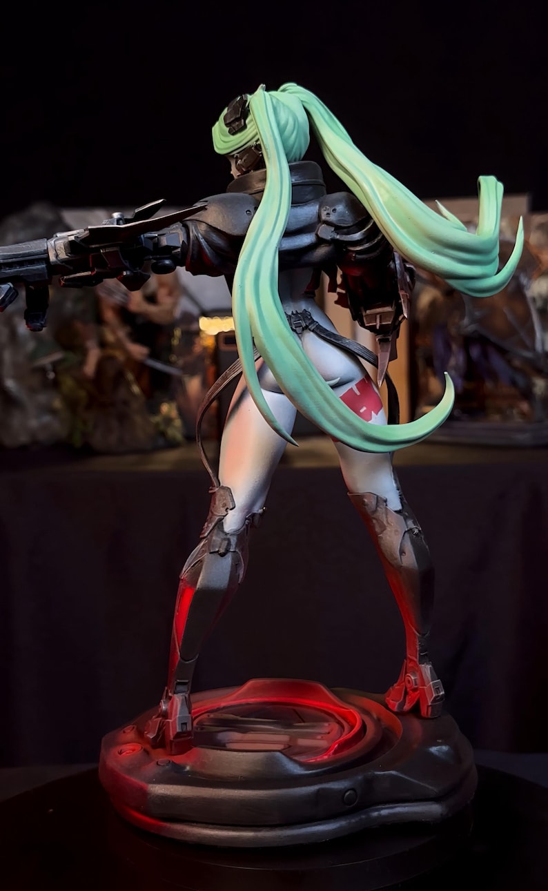 K&ouml;nnte beinhalten: Detaillierte Figur einer weiblichen Figur in einem futuristischen Outfit, die eine Waffe h&auml;lt. Die Figur hat langes, flie&szlig;endes, mintgr&uuml;nes Haar und ist auf einer kreisf&ouml;rmigen Basis mit roter Beleuchtung positioniert. Das Outfit ist haupts&auml;chlich schwarz und grau mit roten Akzenten.