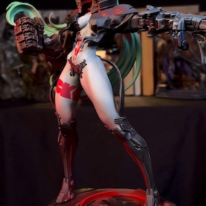 K&ouml;nnte beinhalten: Eine futuristische Figur einer weiblichen Figur mit gr&uuml;nen Haaren und einem wei&szlig;en Gesicht. Sie tr&auml;gt eine schwarz-rote R&uuml;stung und h&auml;lt eine gro&szlig;e Waffe. Die Figur steht auf einem kreisf&ouml;rmigen Sockel mit roter Beleuchtung.
